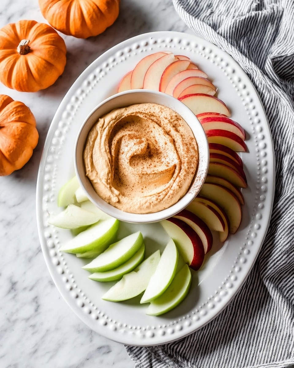 A white bowl filled with light brown creamy dip, swirled with a smooth texture and sprinkled with cinnamon powder on top, with a thin slice of apple placed partially inside the dip. The bowl sits in the middle of a white plate that has a raised dot pattern around the edge. Surrounding the bowl are thin apple slices, some green and some red, neatly arranged around it. The scene is set on a white marbled surface with part of a small orange pumpkin visible in the corner. Photo taken with an iphone --ar 4:5 --v 7
