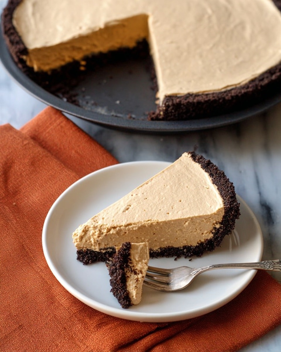 A pie with two layers is shown: the bottom layer is dark brown and crumbly, forming the crust around the edge, while the top layer is thick and light tan with a smooth, creamy texture, spread evenly over the crust. A slice of the pie is placed on a white plate nearby, showing the same two layers and slightly rough edges where it was cut. A silver fork on the plate holds a small bite of the pie, featuring both the dark crust and the tan cream. The background has a white marbled texture, with a folded orange cloth visible beside the plate. photo taken with an iphone --ar 4:5 --v 7
