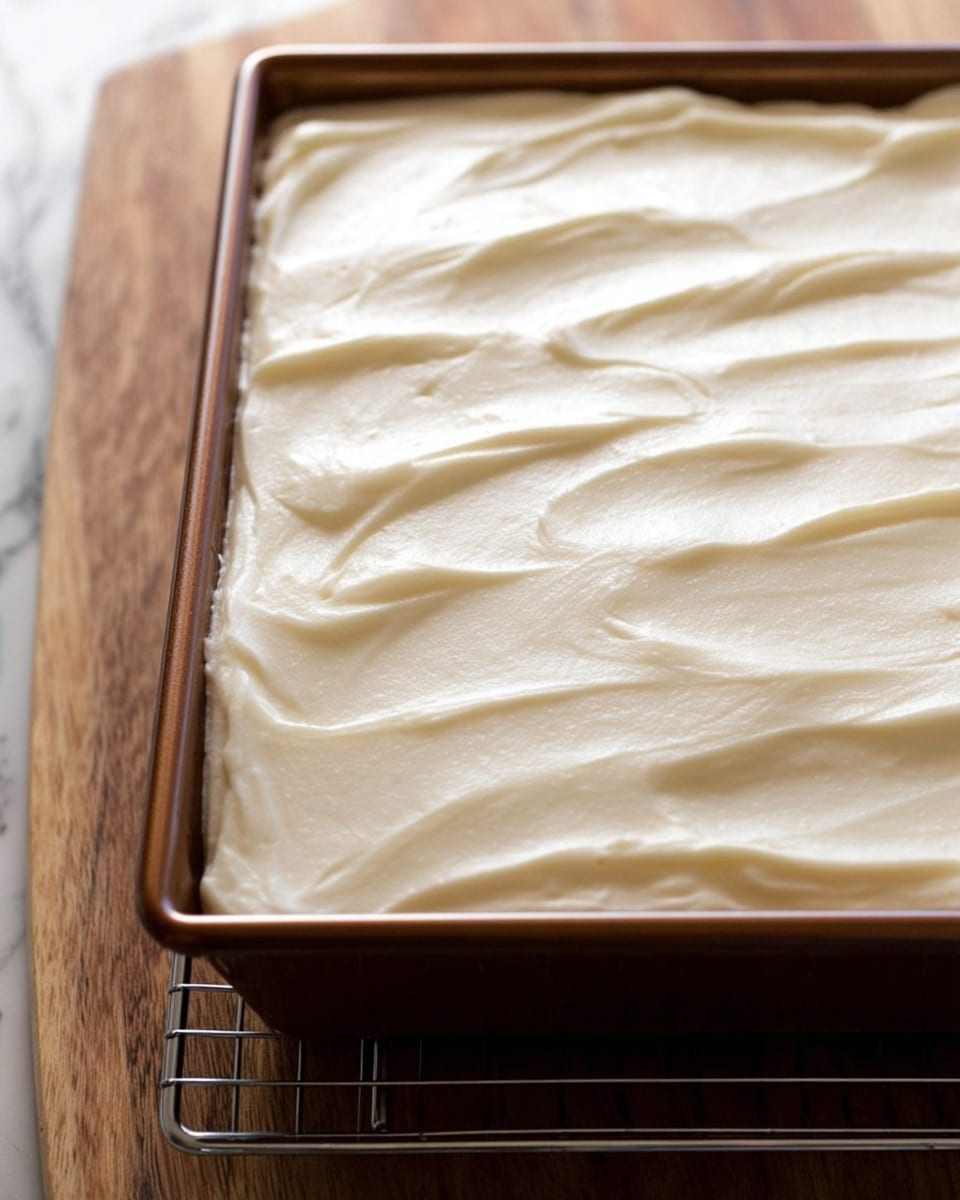A single square piece of moist carrot cake sits on a white round plate, topped with a thick, creamy white frosting layer that looks smooth and soft. The cake layer below is golden brown with visible bits of grated carrot and dark spots, showing a slightly crumbly texture. Next to the cake on the plate is a silver fork resting partially on a green napkin with white and red flower patterns. In the background, a brown baking dish holds the rest of the cake with a large portion missing, showing the same cake and thick white frosting inside. The scene is set on a light wooden surface with a white marbled texture. photo taken with an iphone --ar 4:5 --v 7