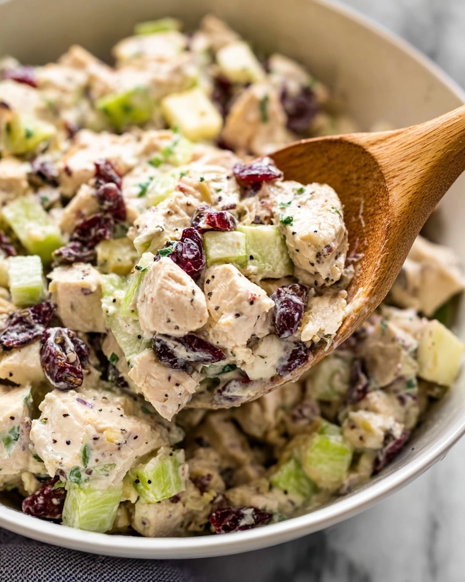 The image shows a close-up of a creamy chicken salad in a white bowl with a wooden spoon. The salad has chunky white pieces of chicken mixed with small chunks of green celery, dark red dried cranberries, and tiny bits of red onion. The creamy dressing coats all the ingredients, giving a soft texture. There are small black pepper flakes sprinkled on top. The bowl is placed on a white marbled textured surface. Photo taken with an iphone --ar 4:5 --v 7