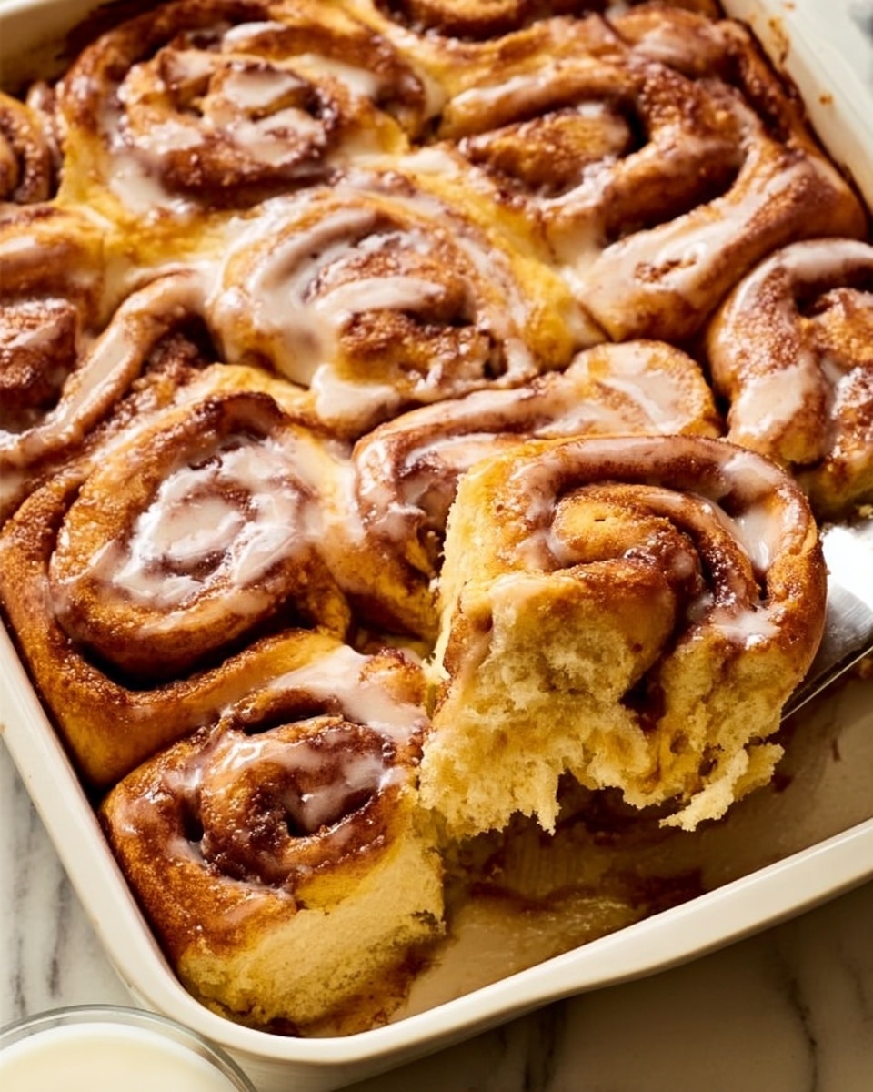 The image shows a white rectangular baking dish filled with tightly packed cinnamon rolls, each with a golden-brown baked top and a light glaze on surface layers that adds a glossy look. The rolls are arranged in a spiral and layered close together, showing thick swirls of cinnamon sugar in the middle of each. One piece is being lifted with a metal spatula, revealing a soft, fluffy inside with a slightly shiny glaze on top. The dish sits on a white marbled surface, and the scene includes a glass of milk partially visible at the edge. photo taken with an iphone --ar 4:5 --v 7