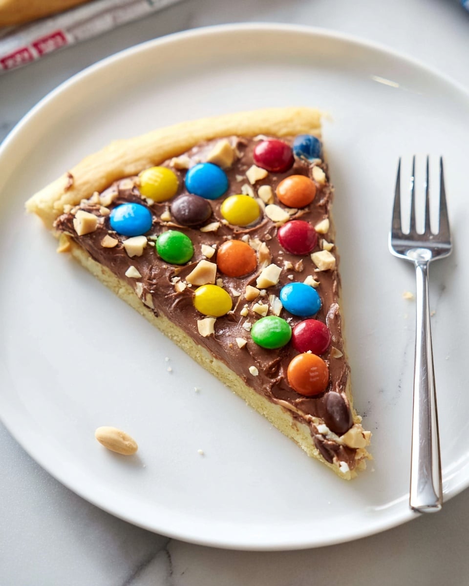 A slice of cookie pizza sits on a clean white round plate, placed on a white marbled surface. The first layer is a light golden cookie base with a soft texture. On top is a smooth, creamy milk chocolate spread covering the whole slice. Scattered over the chocolate are colorful candy-coated chocolates in red, yellow, blue, green, orange, and brown, adding bright pops of color. There are also small pieces of chopped nuts sprinkled unevenly on the slice. A silver fork rests on the right side of the plate. Photo taken with an iphone --ar 4:5 --v 7