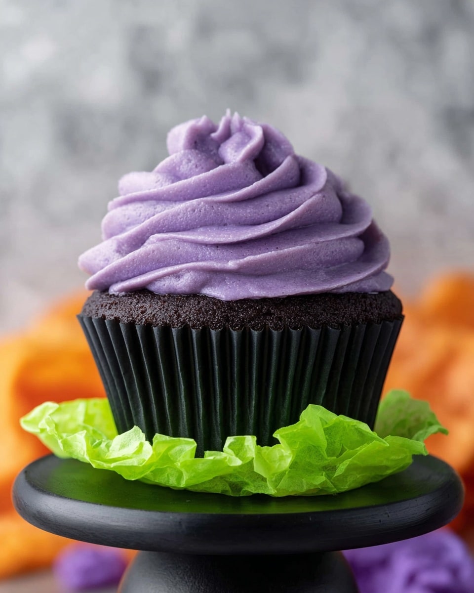 The image shows a dark chocolate cupcake with a black paper liner sitting on bright green crumpled paper, which is placed on top of a smooth black round stand. The cupcake has one thick layer of purple frosting piped in a wavy, swirled texture that rises high above the cupcake, creating a fluffy, soft look. The background is blurred, showing hints of orange and purple colors on a white marbled surface. photo taken with an iphone --ar 4:5 --v 7