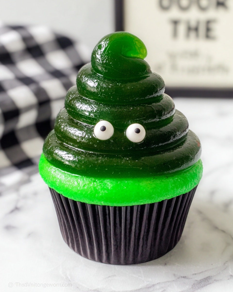 A cupcake in a black paper liner sits on a white marbled surface, topped with a thick base layer of bright green frosting. Above this is a shiny, dark green swirl that looks like soft jelly or gel, shaped into a tall, smooth spiral. Two round white candy eyes with black centers are placed on the front of the green swirl, giving it a playful, character-like look. The background shows a blurry white and black checkered cloth and part of a sign. Photo taken with an iphone --ar 4:5 --v 7