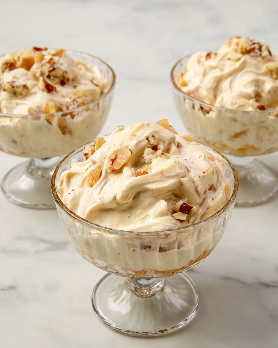 A clear glass bowl sits on a white marbled surface, filled with a creamy mixture that looks thick and smooth. The mixture is off-white with small pieces of light brown and orange chunks scattered inside, giving it a textured look. A bright red spatula is partially immersed in the bowl, the handle resting inside the mixture. The overall look is soft and rich with mixed colors peeking through the cream. photo taken with an iphone --ar 4:5 --v 7