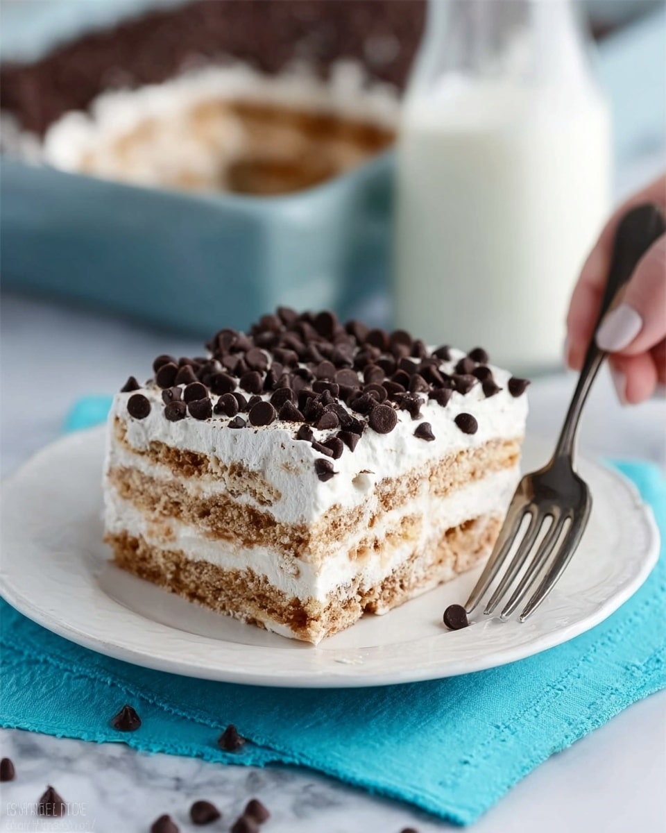 The image shows a white plate with a layered dessert made of four thin golden cake layers separated by smooth white cream layers. The top layer of white cream is decorated with small dark chocolate chips scattered evenly. A silver fork with a piece of dessert on it rests on the plate, and the background features a white marbled surface with a glass of milk and a white pan with more of the dessert slightly blurred behind. photo taken with an iphone --ar 4:5 --v 7