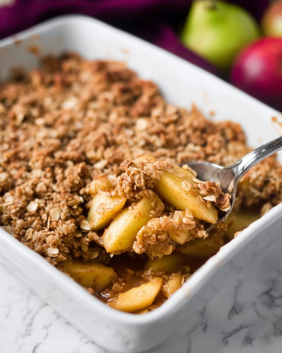The image shows a white rectangular baking dish filled with an apple crumble dessert, featuring two main layers. The bottom layer consists of cooked apple pieces, yellowish in color and coated with a brown cinnamon sauce, visibly soft and juicy. The top layer is a thick, crumbly oat topping that is golden brown and textured with small oats. A silver spoon is scooping a portion from the dish, revealing both layers clearly. The background surface has a white marbled texture with blurred fruits and a purple cloth in the distance. Photo taken with an iphone --ar 4:5 --v 7