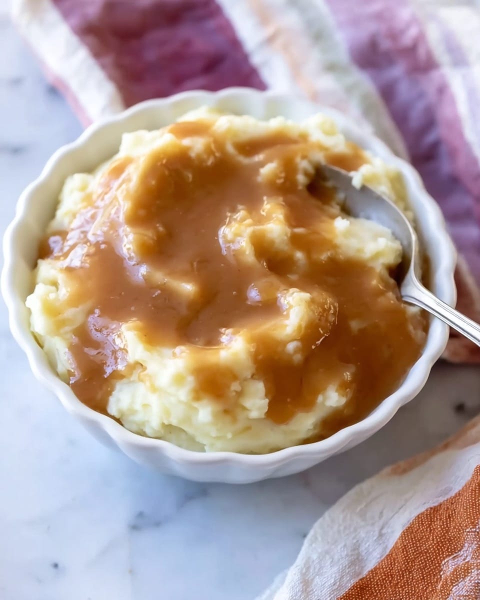 The image shows a white scalloped bowl filled with creamy mashed potatoes as the bottom layer, topped with a thick, smooth brown gravy that covers the potatoes unevenly, creating a mix of light yellow and brown colors with glossy textures. A silver spoon rests partially inside the bowl on the right side, slightly buried in the mashed potatoes. The bowl sits on a white marbled surface with a soft, striped cloth in white, purple, and orange colors beside it. Photo taken with an iphone --ar 4:5 --v 7