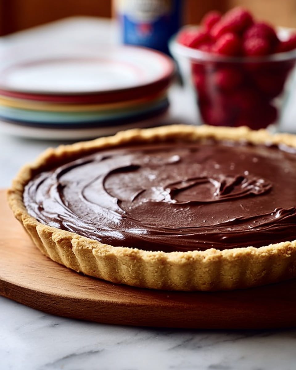 A round tart with a golden-brown crust holds a smooth, thick layer of dark chocolate filling spread evenly with visible swirl patterns on top. The tart sits on a wooden board, and in the background, there is a stack of white plates with colorful rims, a clear container of bright red raspberries, and a blurred can. The surface beneath everything is white marble. The photo is taken close-up with soft natural light, showing a clear texture of the chocolate filling and the crispness of the crust. photo taken with an iphone --ar 4:5 --v 7
