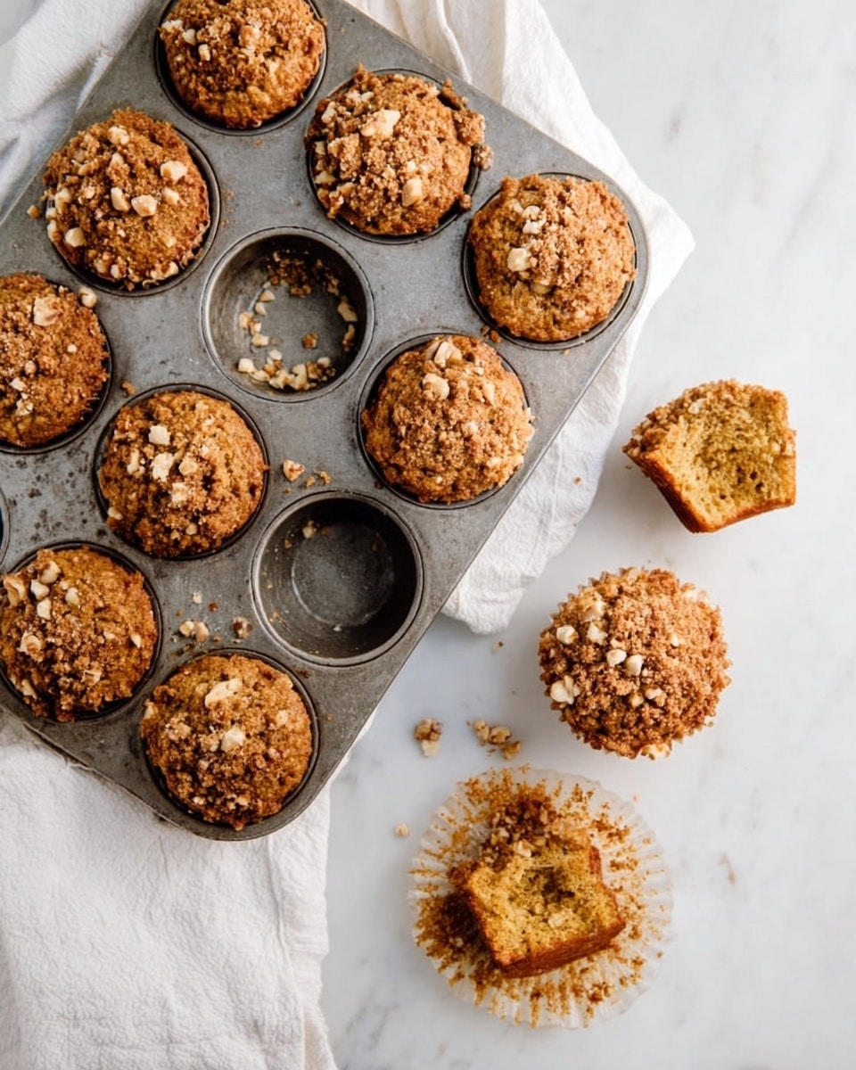 A metal muffin tray resting on a white marbled surface holds eight muffins with a crumbly textured topping made of brown crumbs and small nut pieces. One muffin in the tray is missing, leaving an empty round space with crumbs inside. Two muffins outside the tray sit directly on the white marbled surface; one is whole with the same crumbly topping, and the other is broken in half, revealing a soft, light brown interior. The muffin tray and muffins have a rustic, homemade look, with a white cloth partially visible underneath the tray. Photo taken with an iphone --ar 4:5 --v 7