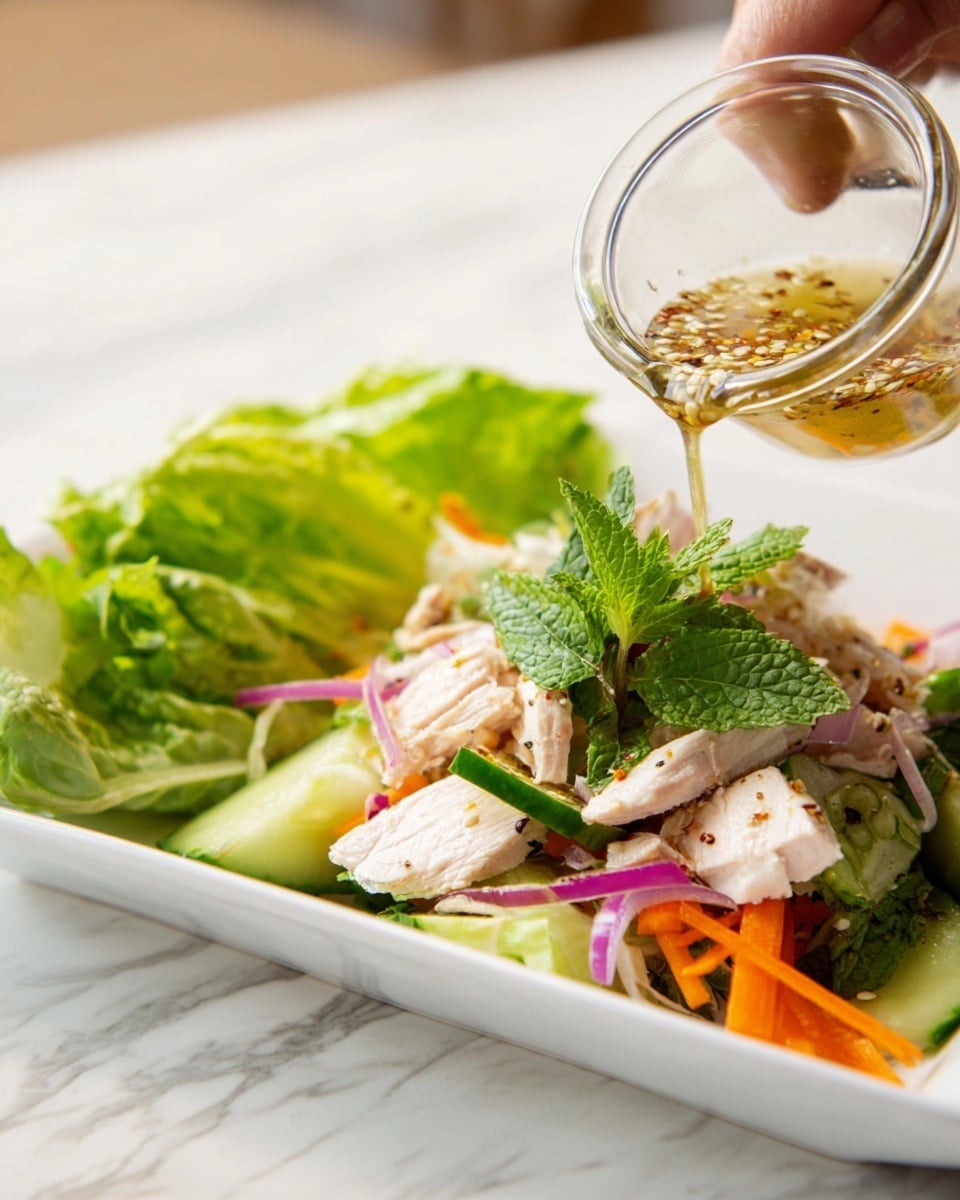 A white square plate holds a fresh salad with several visible layers: at the bottom, there are thin orange carrot strips and sliced green cucumber sticks mixed with purple-red onion slices; above these are pieces of white chicken meat scattered throughout; bright green mint leaves and cilantro sprigs sit on top, adding a fresh look; on one side, there is a small bunch of green romaine lettuce leaves standing upright; above the salad, a woman's hand pours a light-colored dressing with seeds and spices from a clear glass container. The background is a white marbled surface. photo taken with an iphone --ar 4:5 --v 7