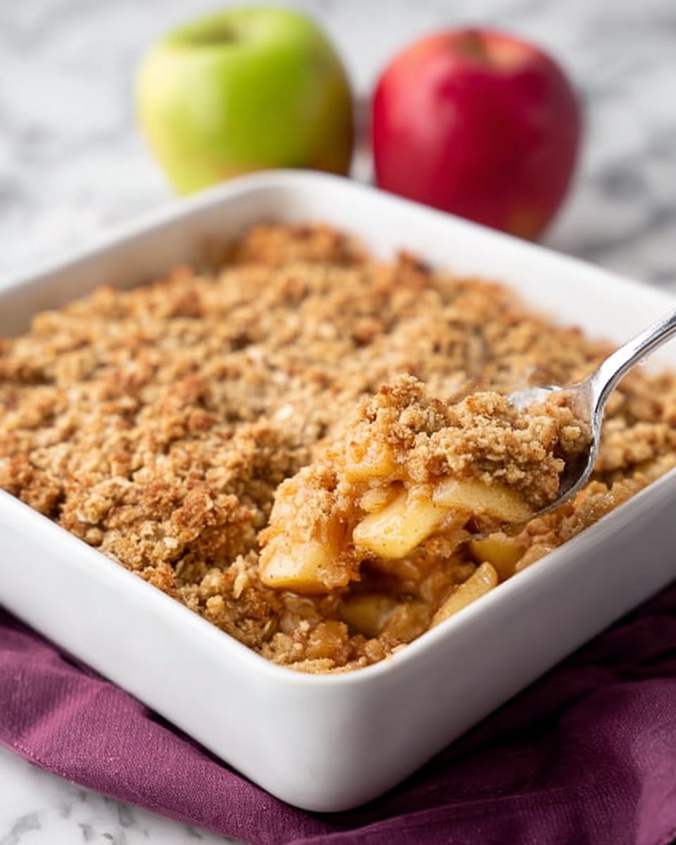A white square baking dish filled with a golden brown apple crumble, featuring a thick crumbly oat topping that covers soft, cooked apple pieces underneath, which are light yellow with hints of cinnamon. A metal spoon is scooping out a portion from the right side, showing the chunky apple filling beneath the textured topping. In the background, a red apple and a green apple sit on a white marbled surface with a purple cloth underneath the dish. Photo taken with an iphone --ar 4:5 --v 7