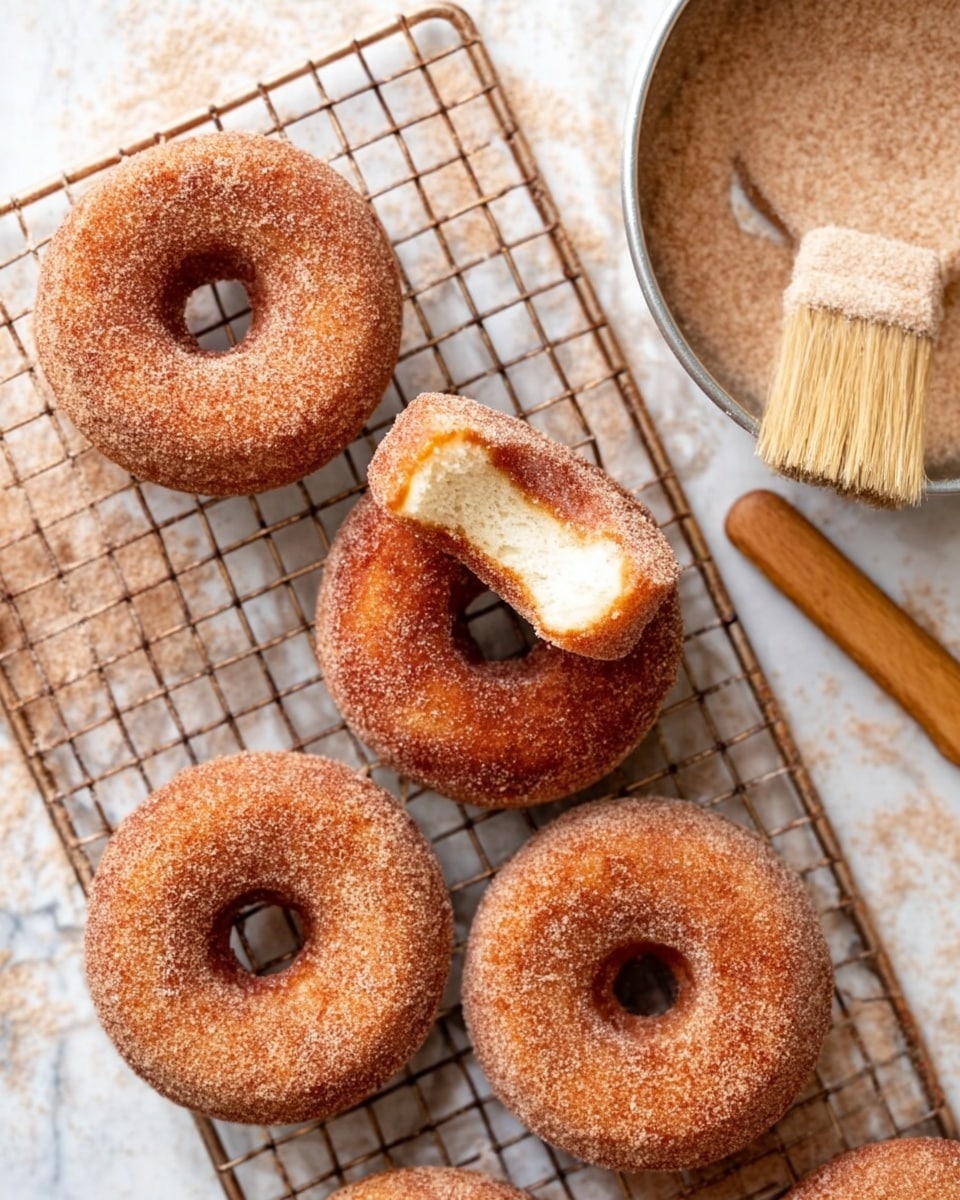 Five cinnamon sugar donuts are arranged on a metal cooling rack over a white marbled surface. Four donuts lay flat, each coated evenly in cinnamon sugar with a textured, slightly rough surface. One donut is held in the center above the others, showing a clear bite taken out of it, revealing a soft, light, fluffy inside with a golden crust. To the top right, a round metal pan filled with cinnamon sugar is partially visible, with a wooden brush resting on its edge with light bristles dusted in cinnamon sugar. Photo taken with an iphone --ar 4:5 --v 7